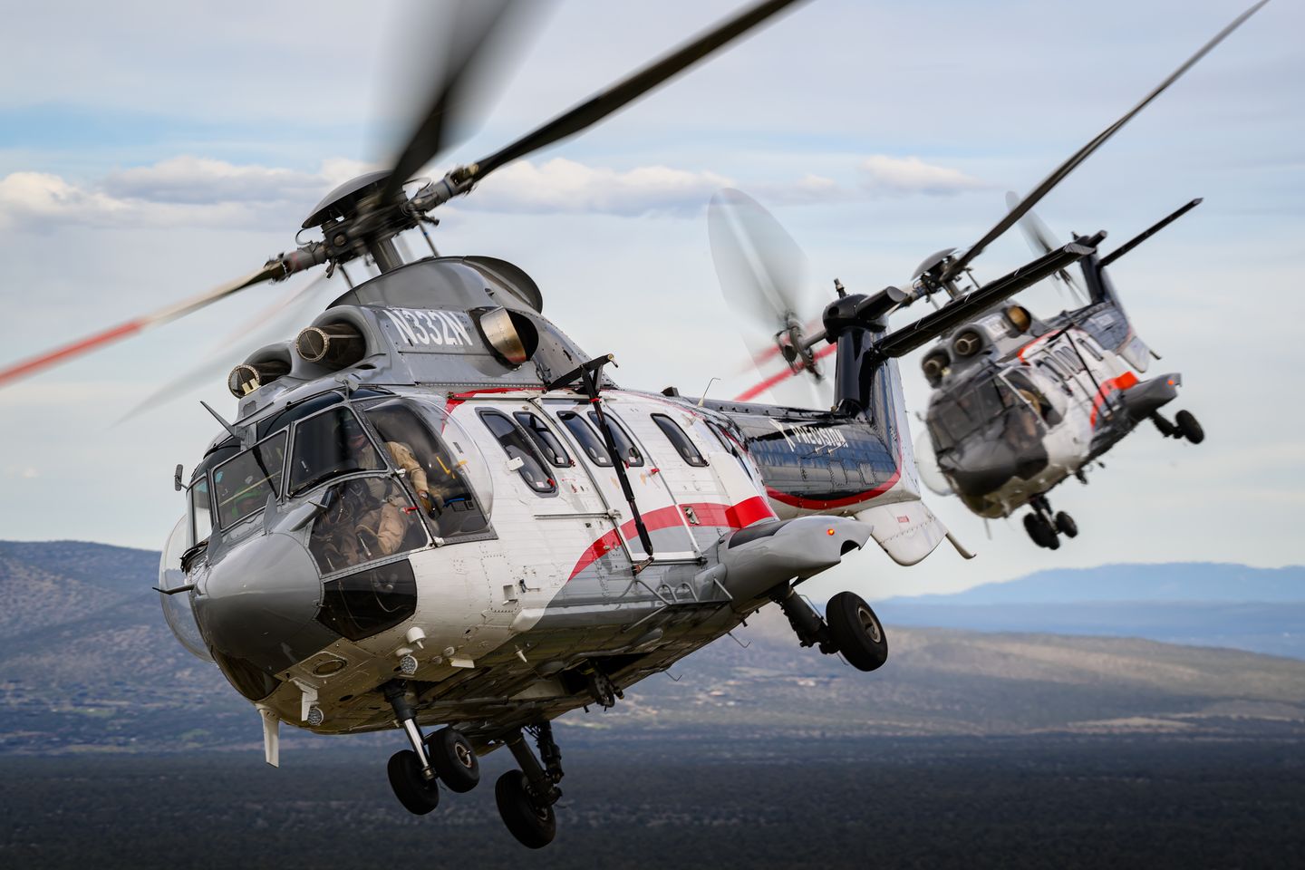 Two Airbus AS332 Super Pumas flying in close formation above high desert terrain.