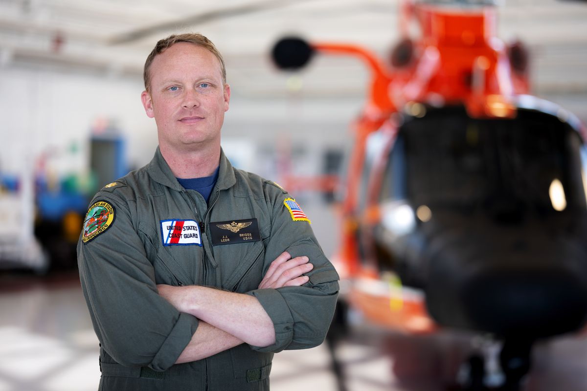 JJ Briggs in a Coast Guard flight suit, arms folded, in a hangar with an MH-65 behind him.